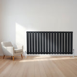 Radiador plano de columna simple negro de 600 x 1190 mm, con válvulas, instalado en una pared junto a una silla moderna en un ambiente luminoso