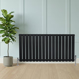 Radiador plano de columna simple negro de 600 x 1190 mm, con válvulas, colocado frente a una pared y una planta decorativa