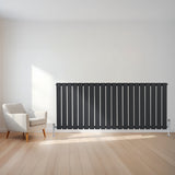 Radiador plano de columna simple en gris antracita, con válvulas, instalado en una pared blanca junto a una silla moderna de madera clara