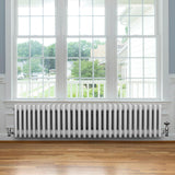 Radiador tradicional de 4 columnas en color blanco, ubicado bajo una ventana, con un diseño elegante y acabado brillante sobre un suelo de madera clara