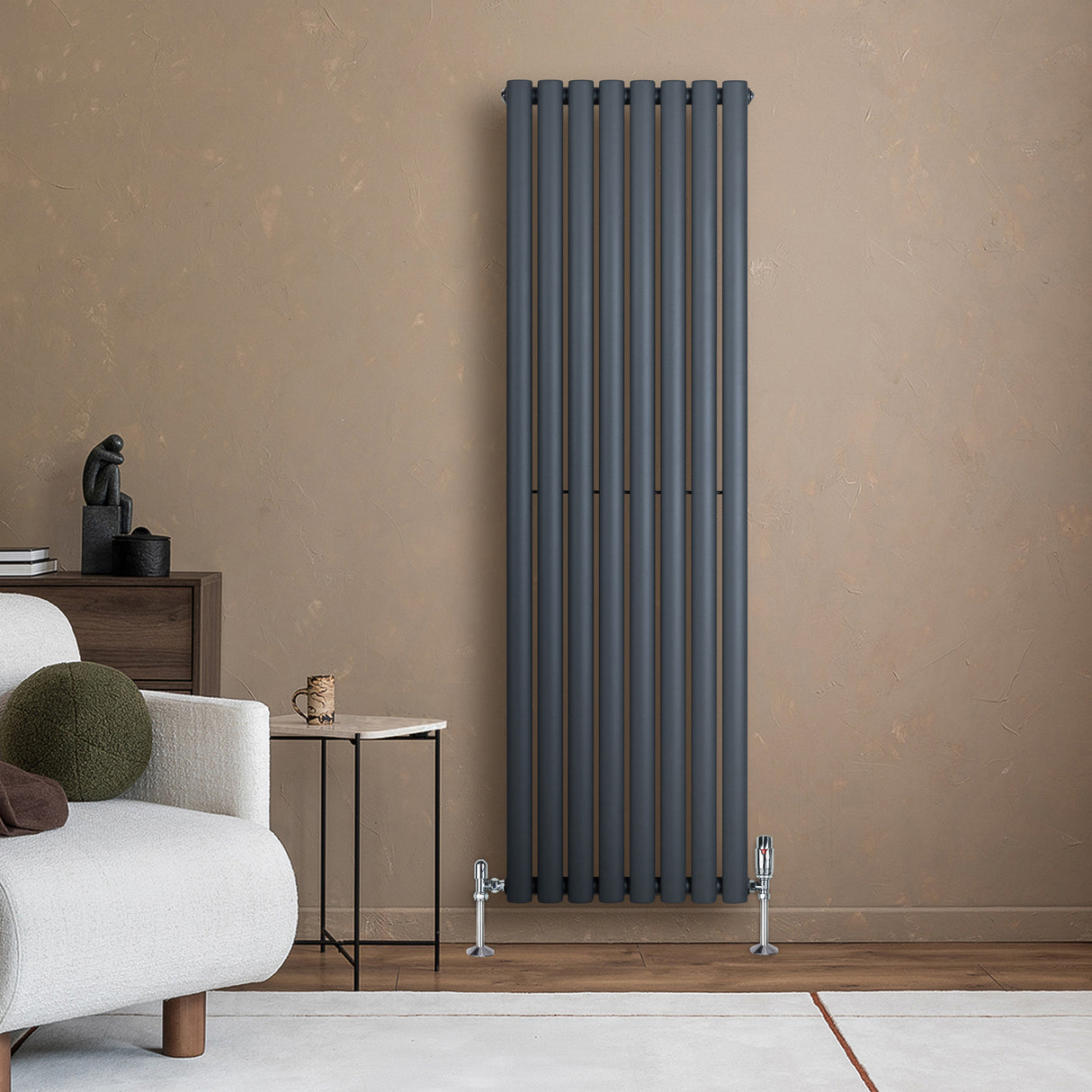 Radiador de columna ovalado en gris antracita, con un diseño vertical y acabado mate, instalado en una pared beige junto a un sofá moderno