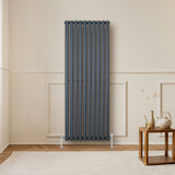 Radiador de columna ovalado en gris antracita, con diseño vertical y acabado mate, instalado en una pared clara junto a una mesa de madera