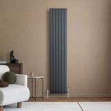 Radiador de columna ovalado en gris antracita, con diseño moderno y acabado mate, instalado en pared beige junto a un sofá claro