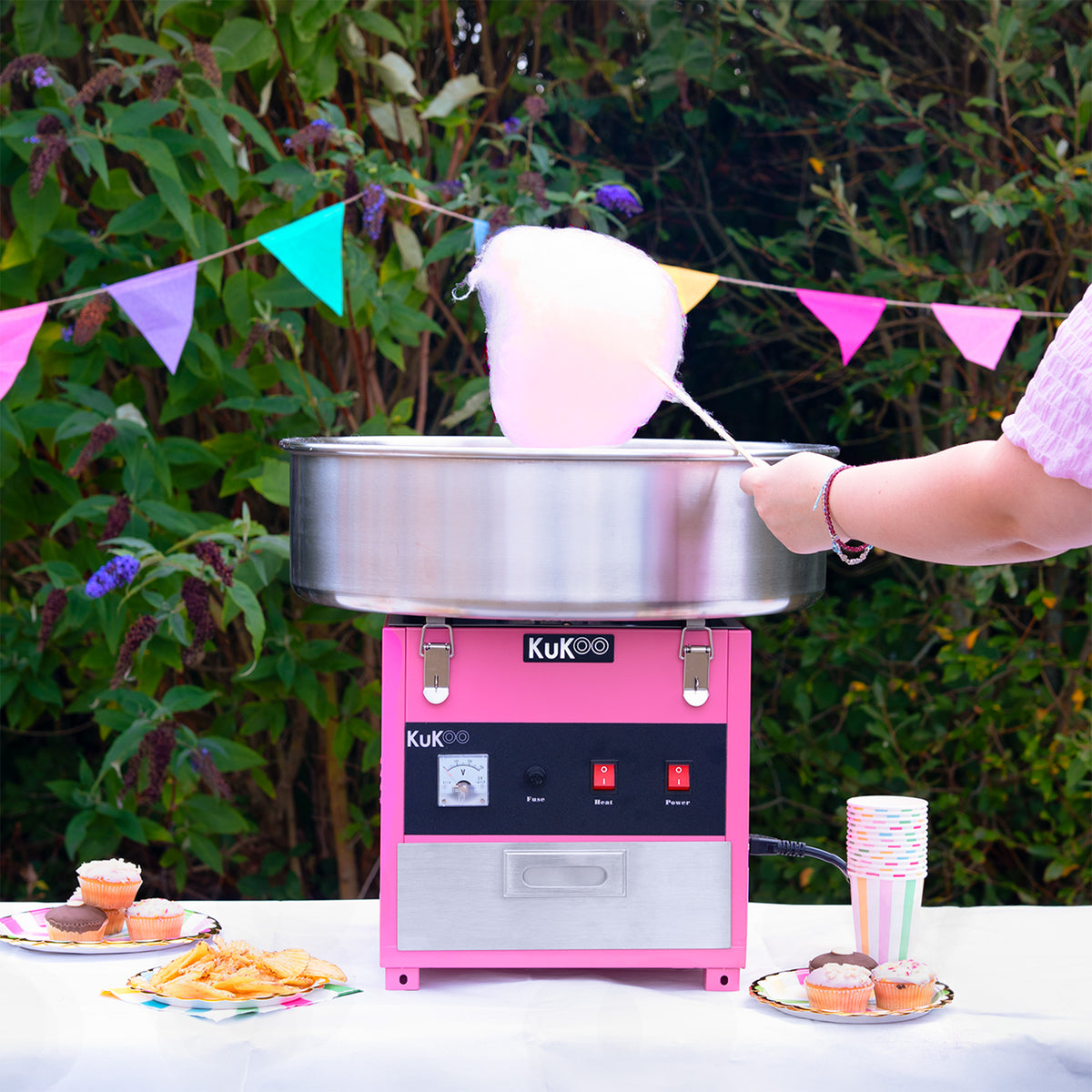 Máquina de algodón de azúcar KuKoo de color rosa, con un tazón de acero inoxidable, produciendo nubes de algodón de azúcar en un ambiente festivo