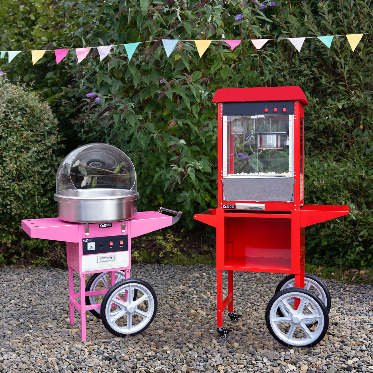 Máquina de palomitas rosa y carrito de algodón de azúcar rojo, ambos con ruedas, en un entorno al aire libre con banderines decorativos