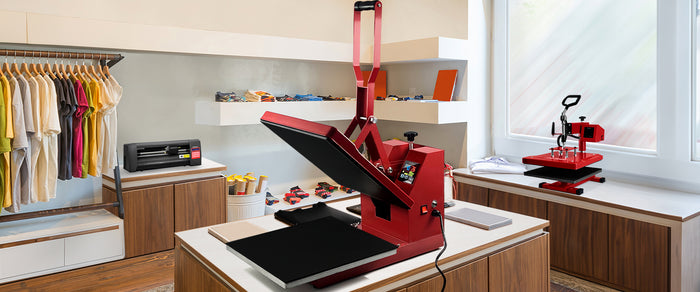 Prensa térmica roja sobre una mesa negra, rodeada de ropa y herramientas de diseño en un entorno luminoso y moderno