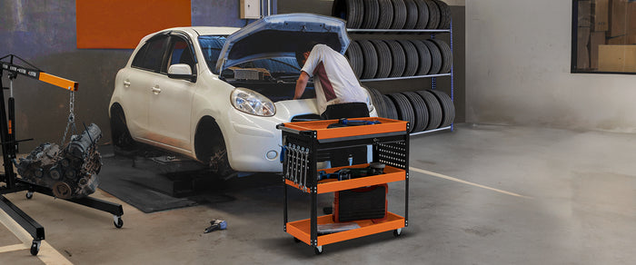 Carro de herramientas naranja y negro en un taller, junto a un coche blanco con el capó abierto y un mecánico trabajando en el motor