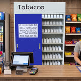 Puerta azul con letrero que prohíbe la venta de tabaco a menores de 18 años, fondo de estantes con productos y mostrador de caja